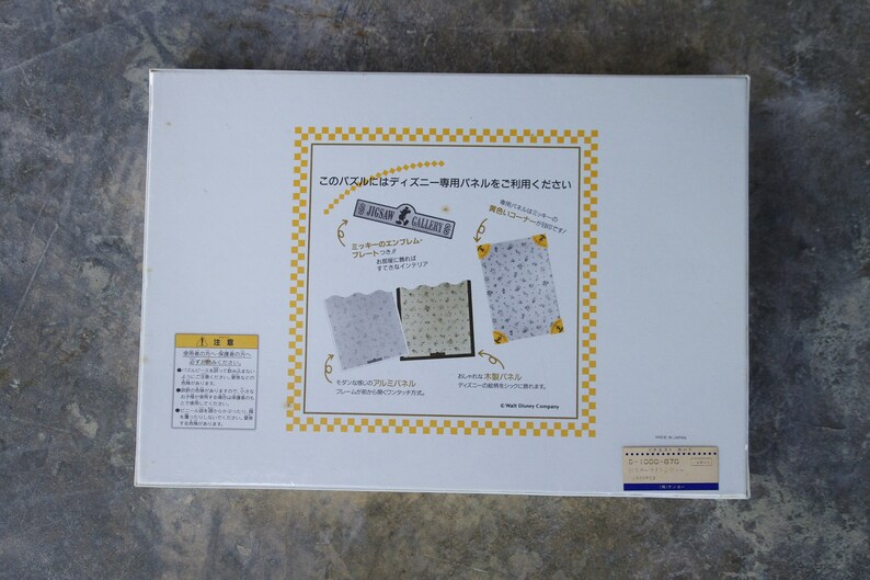 May include: White box with a yellow and black checkered border. The box features a picture of a jigsaw puzzle with a Mickey Mouse design. The text on the box reads "Jigsaw Gallery" and "This puzzle uses Disney-exclusive panels".