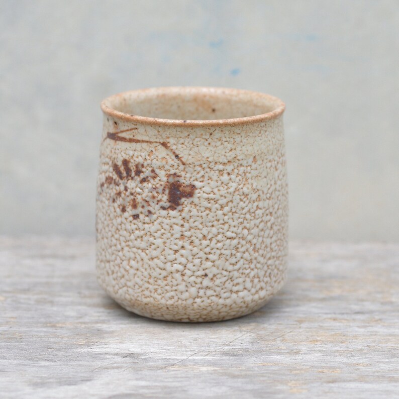 Puede incluir: Una taza peque&ntilde;a de cer&aacute;mica blanca con una superficie texturizada y marcas marrones. La taza est&aacute; sobre una superficie de madera clara.