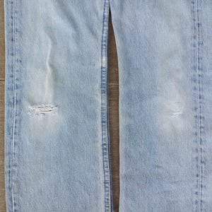 Puede incluir: Vaqueros de mezclilla azul claro con detalles desgastados, incluyendo una rodilla rasgada. Los vaqueros tienen un dise&ntilde;o cl&aacute;sico de cinco bolsillos y un lavado descolorido, mostrando signos de desgaste. La imagen muestra la parte delantera de los vaqueros.