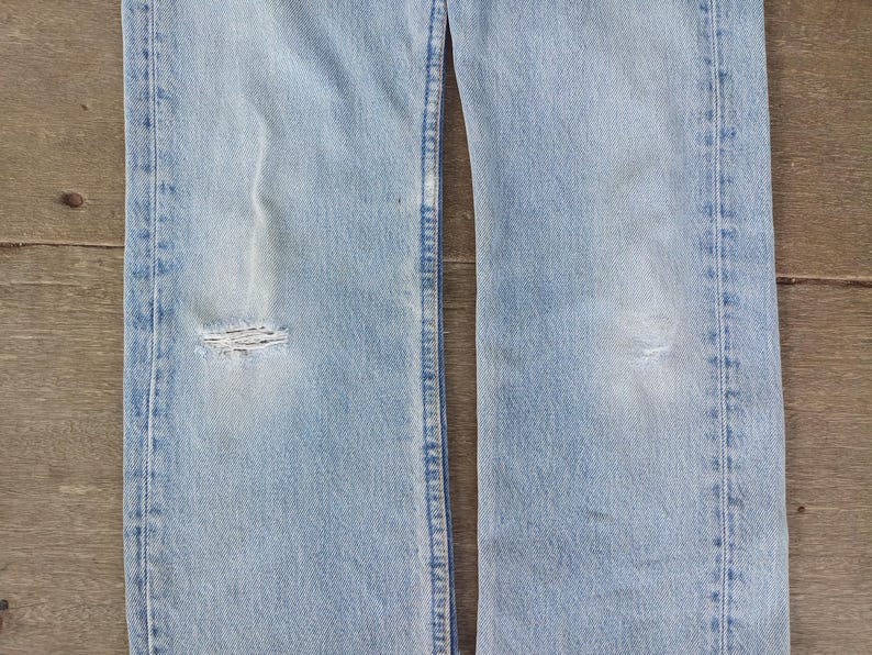 Puede incluir: Vaqueros de mezclilla azul claro con detalles desgastados, incluyendo una rodilla rasgada. Los vaqueros tienen un dise&ntilde;o cl&aacute;sico de cinco bolsillos y un lavado descolorido, mostrando signos de desgaste. La imagen muestra la parte delantera de los vaqueros.