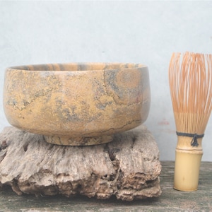 Puede incluir: Un cuenco de matcha de piedra marrón y amarillo con un batidor de bambú sobre una superficie de madera.