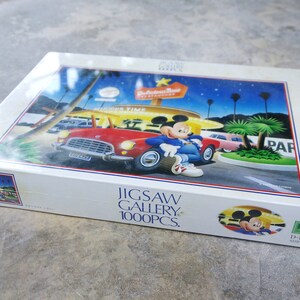 May include: A 1000 piece jigsaw puzzle featuring Mickey Mouse in a red car in front of a diner with a sign that says "Hollywood Time" and "Restaurant". The puzzle box is white with blue and red accents. The puzzle is made by Disney.