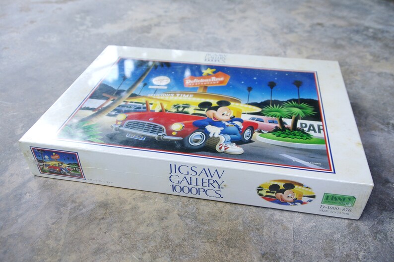 May include: A 1000 piece jigsaw puzzle featuring Mickey Mouse in a red car in front of a diner with a sign that says "Hollywood Time" and "Restaurant". The puzzle box is white with blue and red accents. The puzzle is made by Disney.