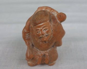以下が含まれることがあります： 背中に荷物を背負った男性の小さな茶色の粘土の置物。男性は、口ひげとあごひげのある詳細な顔をしています。