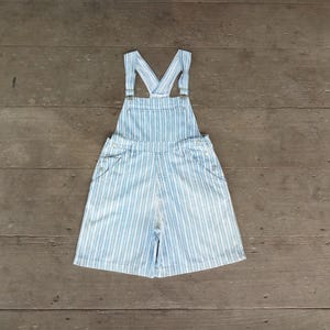 May include: Light blue and white striped overall shorts with adjustable straps. The shorts have two front pockets and two back pockets. The overall shorts are made of a lightweight denim material and are laid flat on a wooden surface.