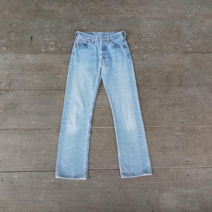 Puede incluir: Vaqueros de mezclilla azul claro con bragueta de botones y dise&ntilde;o cl&aacute;sico de cinco bolsillos. Los vaqueros tienen un lavado descolorido y una rodilla desgastada. Los vaqueros est&aacute;n planos sobre una superficie de madera.