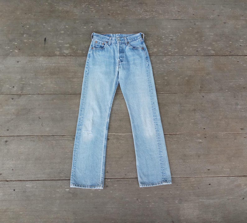 Puede incluir: Vaqueros de mezclilla azul claro con bragueta de botones y dise&ntilde;o cl&aacute;sico de cinco bolsillos. Los vaqueros tienen un lavado descolorido y una rodilla desgastada. Los vaqueros est&aacute;n planos sobre una superficie de madera.