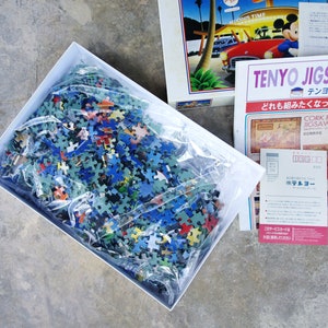 May include: A white box containing a plastic bag filled with a jigsaw puzzle pieces. The pieces are various colors, including blue, green, red, and yellow. The box is sitting on a gray concrete surface. The text on the box reads "Tenyo Jigsaw Puzzle".