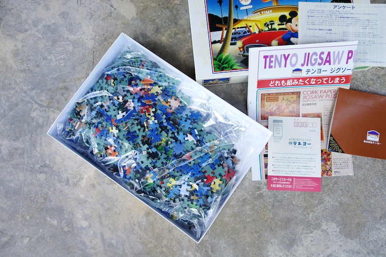 May include: A white box containing a plastic bag filled with a jigsaw puzzle pieces. The pieces are various colors, including blue, green, red, and yellow. The box is sitting on a gray concrete surface. The text on the box reads "Tenyo Jigsaw Puzzle".