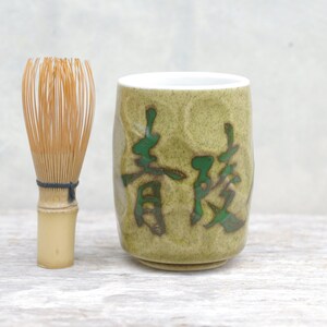 May include: A green ceramic teacup with Japanese characters and a bamboo whisk. The teacup has a textured surface and a light green glaze. The whisk is made of natural bamboo.