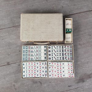 Könnte beinhalten: Ein Mahjong-Set in einem Holzkasten. Die Steine sind weiß mit schwarzen und roten Schriftzeichen. Der Kasten ist braun mit einem weißen Innenraum.