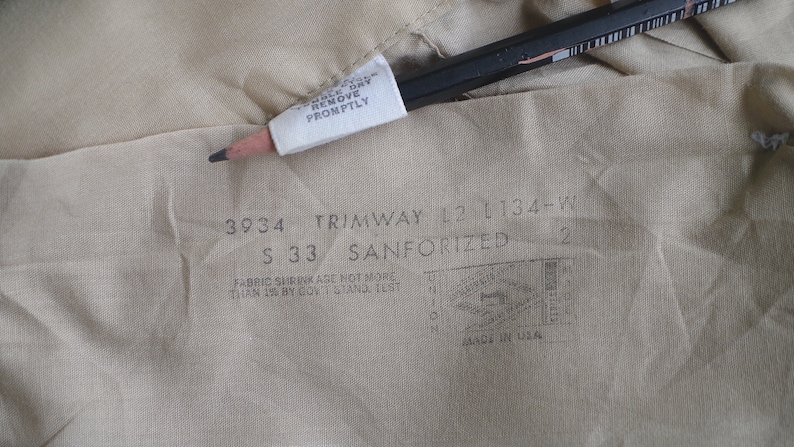 May include: A close-up of a beige fabric label with the text "3934 TRIMWAY 12 1134-W S 33 SANFORIZED 2" and "FABRIC SHRINKAGE NOT MORE THAN 1% BY GOV'T STAND TEST MADE IN USA".