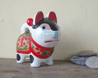 May include: A white, black, and red wooden cat figurine with a floral design. The cat has a red collar and black eyes. The figurine is standing on a wooden surface.
