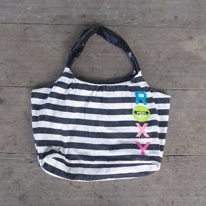 May include: A black and white striped tote bag with a black leather handle. The bag has the word "ROXY" printed on the front in colourful letters.