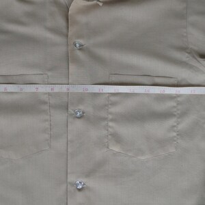 May include: A beige button-down shirt with two chest pockets and a tape measure across the front. The tape measure reads 21 inches.