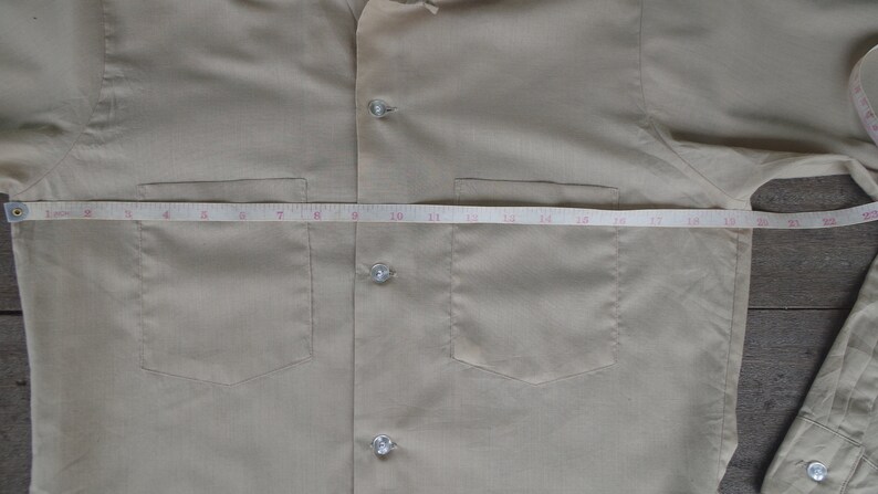 May include: A beige button-down shirt with two chest pockets and a tape measure across the front. The tape measure reads 21 inches.