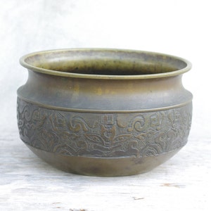 May include: A round, bronze planter with an intricate, embossed design. The planter has a wide, rounded base and a slightly flared lip.