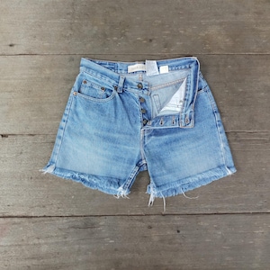Puede incluir: Shorts vaqueros azul claro con bragueta de botones y dobladillo deshilachado. Los shorts tienen un diseño clásico de cinco bolsillos y están colocados sobre una superficie de madera. Los shorts son de mezclilla de lavado claro.