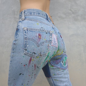 Könnte beinhalten: Hellblaue Jeans aus Denim mit Farbspritzern und bunten Patches. Die Jeans haben eine Gesäßtasche mit einem weißen Etikett.