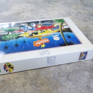 May include: A white box with a colorful image of Mickey Mouse and friends in front of a building with the text "Disneyland" on it. The box is for a 1000 piece jigsaw puzzle.