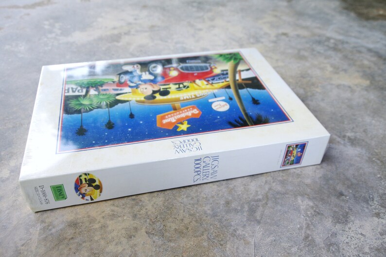 May include: A white box with a colorful image of Mickey Mouse and friends in front of a building with the text "Disneyland" on it. The box is for a 1000 piece jigsaw puzzle.