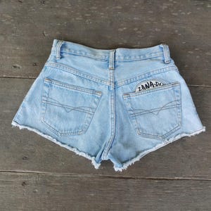 Puede incluir: Shorts vaqueros azul claro con dobladillo deshilachado. Los shorts tienen dos bolsillos traseros y un pequeño parche con el texto "ZANA-DI". Colocados sobre una superficie de madera.
