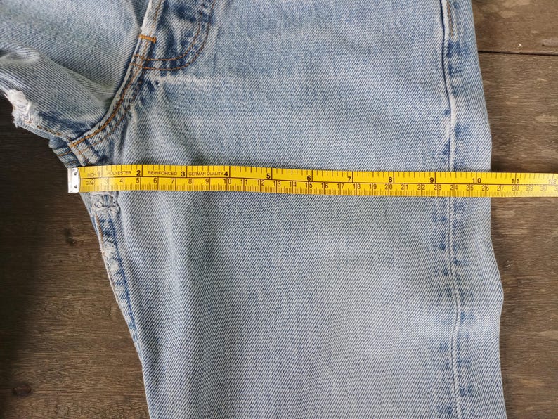Puede incluir: Vaqueros de mezclilla azul claro con una cinta m&eacute;trica amarilla que muestra una medida de aproximadamente 25 cm. Los vaqueros tienen un aspecto desgastado con algunos deshilachados en los bordes. Los vaqueros est&aacute;n sobre una superficie de madera.