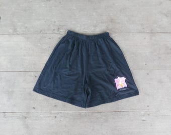 May include: Dark navy blue shorts with an elastic waistband. A pink and white floral design with the word "Hawaii" is printed on the lower right leg. The shorts are made of a soft, lightweight material.