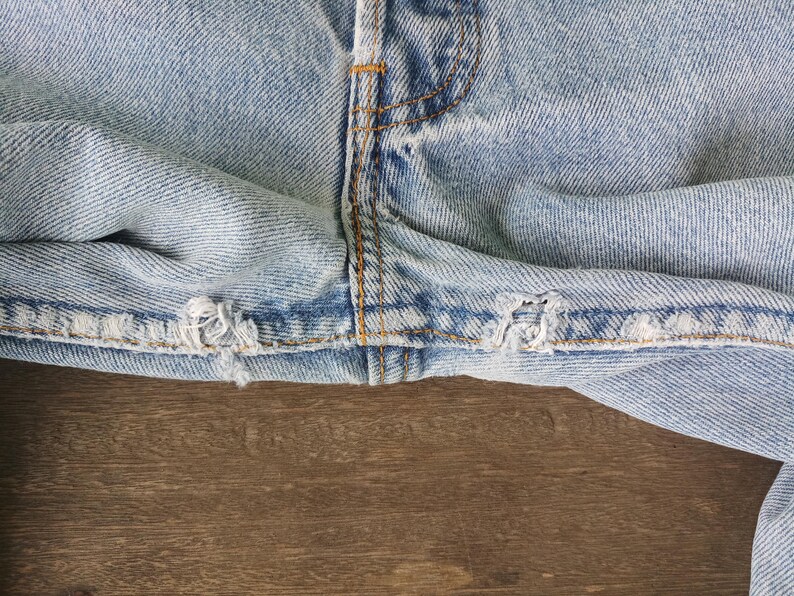 Puede incluir: Primer plano de unos vaqueros de mezclilla azul claro, que muestran el dobladillo desgastado y los detalles de las costuras. Los vaqueros tienen un aspecto desgastado con deshilachados y roturas visibles a lo largo del borde inferior. Las costuras est&aacute;n reforzadas con hilo amarillo en contraste.
