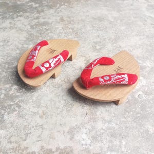 May include: A pair of traditional Japanese geta sandals. The wooden platforms are light brown with a curved shape and small wooden supports. The red fabric straps are decorated with a silver floral pattern.