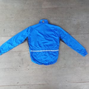 May include: A bright blue, long-sleeved jacket with a high collar and a reflective stripe across the lower back. The jacket is laid flat on a wooden surface, showcasing its design and features. The jacket is a lightweight garment.