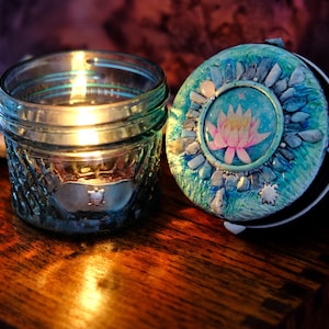 Puede incluir: Un portavelas de cristal transparente con un patrón de diamantes texturizado y un pequeño emblema de tortuga plateado, acompañado de una tapa circular con una ilustración central de una flor de loto rosa rodeada de acentos de piedra azul y verde.