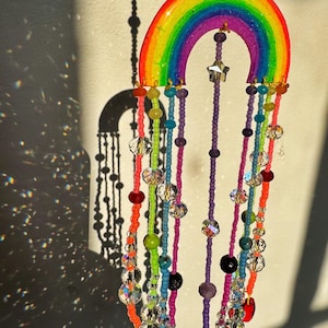 May include: A colorful rainbow-shaped suncatcher with beaded strands. The rainbow features vibrant hues of red, orange, yellow, green, blue, and purple. Strands of beads in various colors and crystal accents hang below, creating a decorative piece.