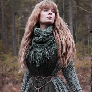 Puede incluir: Una mujer con un vestido verde oscuro con un chal y bufanda de ganchillo a juego. El vestido tiene una falda larga y un corpiño ajustado, ceñido a la cintura con un cinturón de cuerda marrón. El chal y la bufanda son de un verde más claro con flecos.