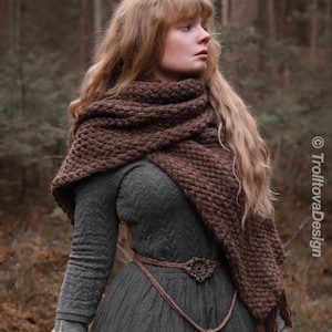 May include: A woman wearing a dark green dress, brown shawl, and a rope belt with a decorative buckle. The shawl is a dark brown, knitted design. The dress has long sleeves and a textured pattern. The background is a blurred forest.