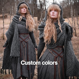 May include: A woman wearing a long grey dress with a brown leather belt and a grey knitted hooded scarf with tassels. The scarf is worn as a shawl and has a braided design. The woman has long blonde hair and is standing in a woodland setting. The text "Custom colours" is visible in the image.