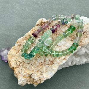 Puede incluir: Una pulsera de cuentas hecha de pequeñas cuentas de fluorita translúcidas en forma de disco. Las cuentas muestran un degradado de colores que va desde el morado intenso y lavanda hasta varios tonos de verde claro y oscuro, ensartadas en un cordón elástico blanco.