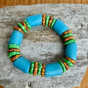 May include: A colourful bracelet with large, light blue cylindrical beads and smaller, round beads in green, yellow, and brown. The bracelet is displayed on a weathered wooden surface.