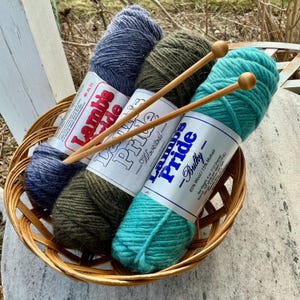 May include: A wicker basket holds three skeins of Lamb's Pride yarn in blue, olive green, and teal, with two wooden knitting needles. The yarn labels are visible. The basket sits on a weathered surface.