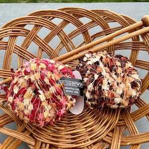 May include: Two colorful yarn balls in shades of red, pink, and brown sit in a woven basket with a pair of wooden knitting needles. The yarn balls have a textured, ribbon-like appearance. The basket is made of woven wicker.