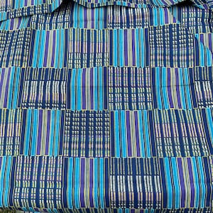 May include: Close-up of a fabric with a geometric pattern. The design features vertical stripes in shades of blue, turquoise, and purple, interspersed with sections of white and yellow. The fabric has a textured appearance, suggesting a woven material.