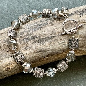 May include: Silver-toned bracelet featuring square and faceted beads. The square beads have a swirled design, and the faceted beads are clear. The bracelet has a toggle clasp. The jewelry is displayed on a piece of wood.