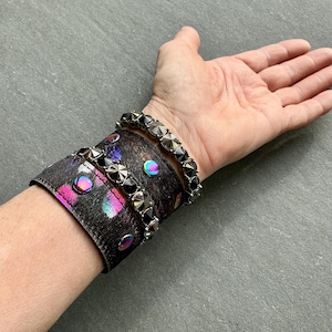 Puede incluir: Un brazalete de cuero negro con un diseño abstracto colorido y tachuelas plateadas. También se ve un brazalete plateado a juego con tachuelas. El brazalete presenta detalles iridiscentes.