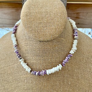 Könnte beinhalten: Eine Halskette aus weißen und lila Muschelsplittern. Die Halskette wird auf einem beigefarbenen Schaufensterpuppenkopf präsentiert. Die Muschelteile sind unregelmäßig geformt und zu einer Halskette aufgereiht.