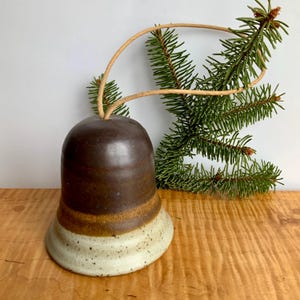 May include: A handcrafted ceramic bell with a brown top, a tan band, and a speckled white base. A natural twine loop is attached for hanging. The bell is displayed with a sprig of green pine needles, suggesting a holiday theme.