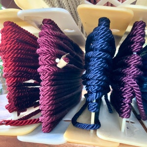 May include: Close-up of three spools of thick, braided cord. The cords are burgundy, navy blue, and a darker shade of burgundy. The cords are wound around white plastic spools.