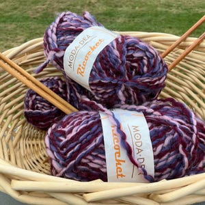 Pode incluir: Uma cesta de vime contém dois novelos de lã Moda Dea Ricochet em tons de roxo, rosa e azul. Duas agulhas de tricô de madeira estão na cesta. A lã é grossa e texturizada, perfeita para projetos de tricô ou crochê.