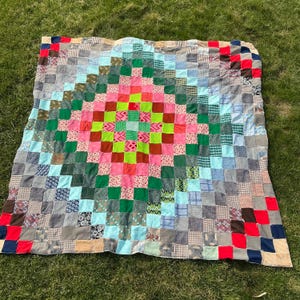 May include: A colorful patchwork quilt with a central diamond pattern. The quilt features squares of various colors, including pink, green, blue, and gray. The quilt is laid out on a grassy surface, showcasing its handmade quality and vibrant design.