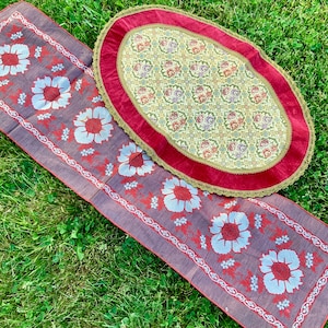 May include: Two floral patterned table runners. One is a round oval shape with a red border and a cream background. The other is a rectangular runner with a brown background and a red border. Both runners have a floral pattern with red, white, and green flowers.
