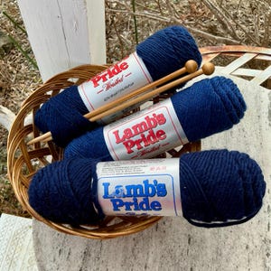 May include: Three navy blue yarn skeins labeled "Lamb's Pride" and "Pride Worsted" are nestled in a woven basket. Two wooden knitting needles rest across the yarn. The yarn is 85% wool and 15% mohair.